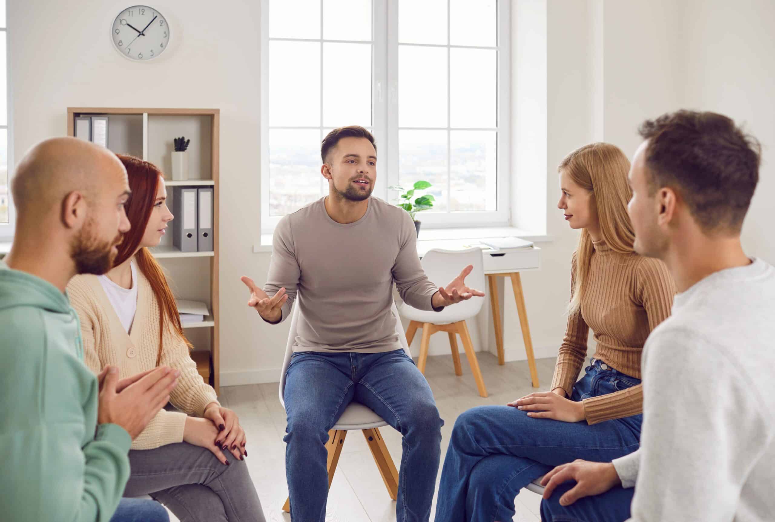 A diverse group of adults participating in a clinical group therapy session at a mental health treatment facility, emphasizing peer support and open communication in a professional, light-filled environment.